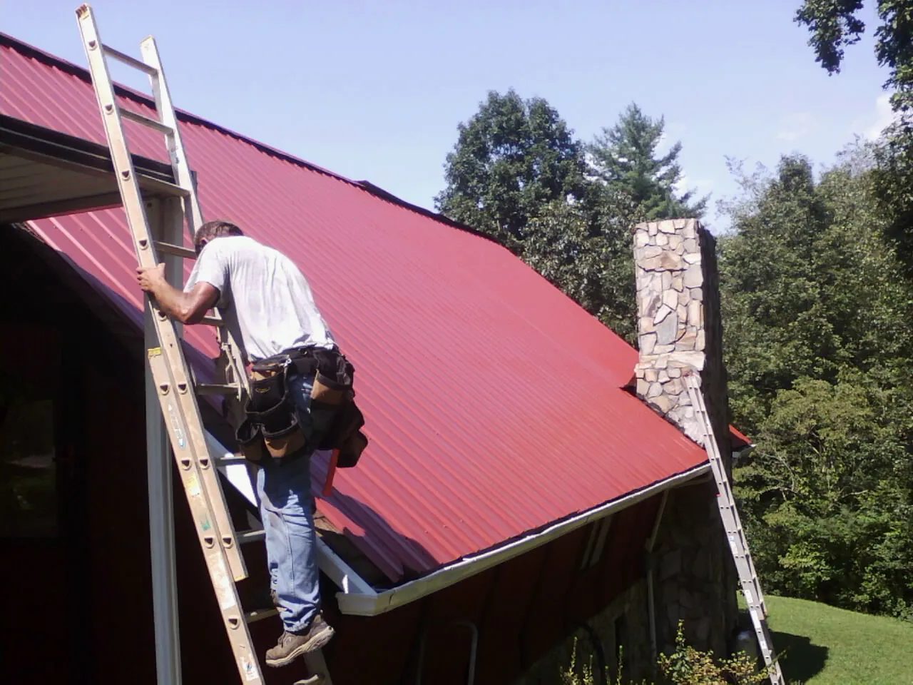 Roofer on a ladder beside a red metal roof and stone chimney — Soffit & Fascia Repair by Roofing Brothers in Virginia Beach